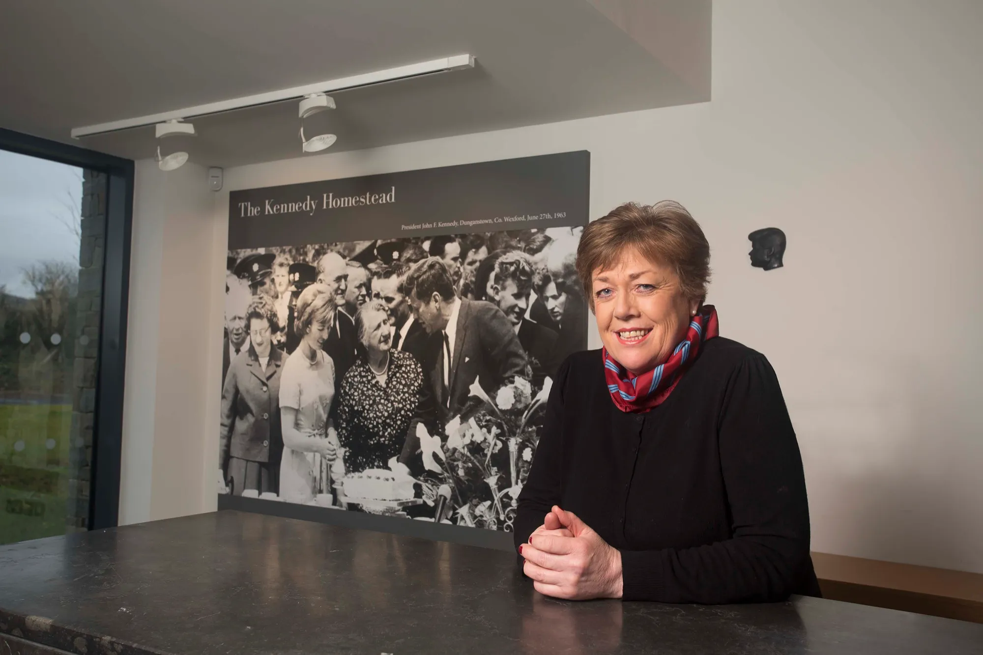 A member of the homestead team smiling at the reception desk, in front of a large black-and-white photograph of the 1963 visit.