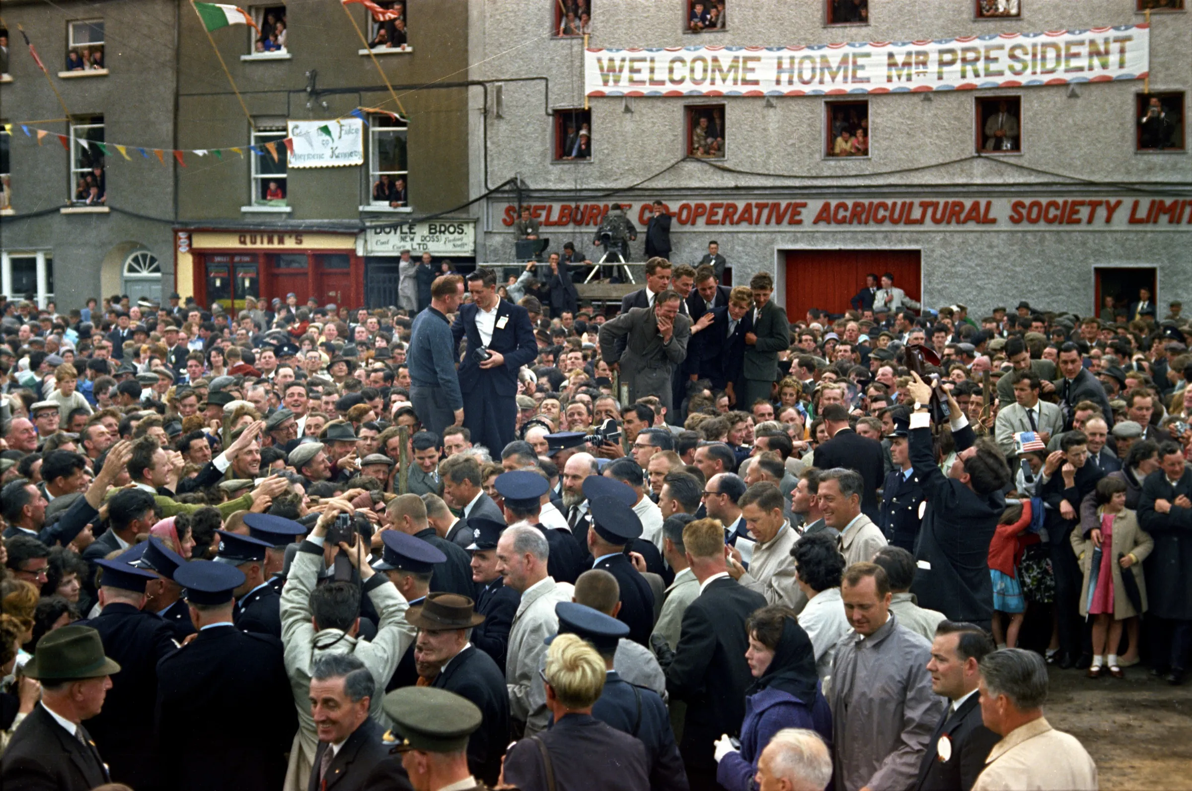 A huge crowd on the New Ross quay, packed in front of a stone building hung with a banner reading Welcome Home Mr President, with Gardaí in the foreground.
