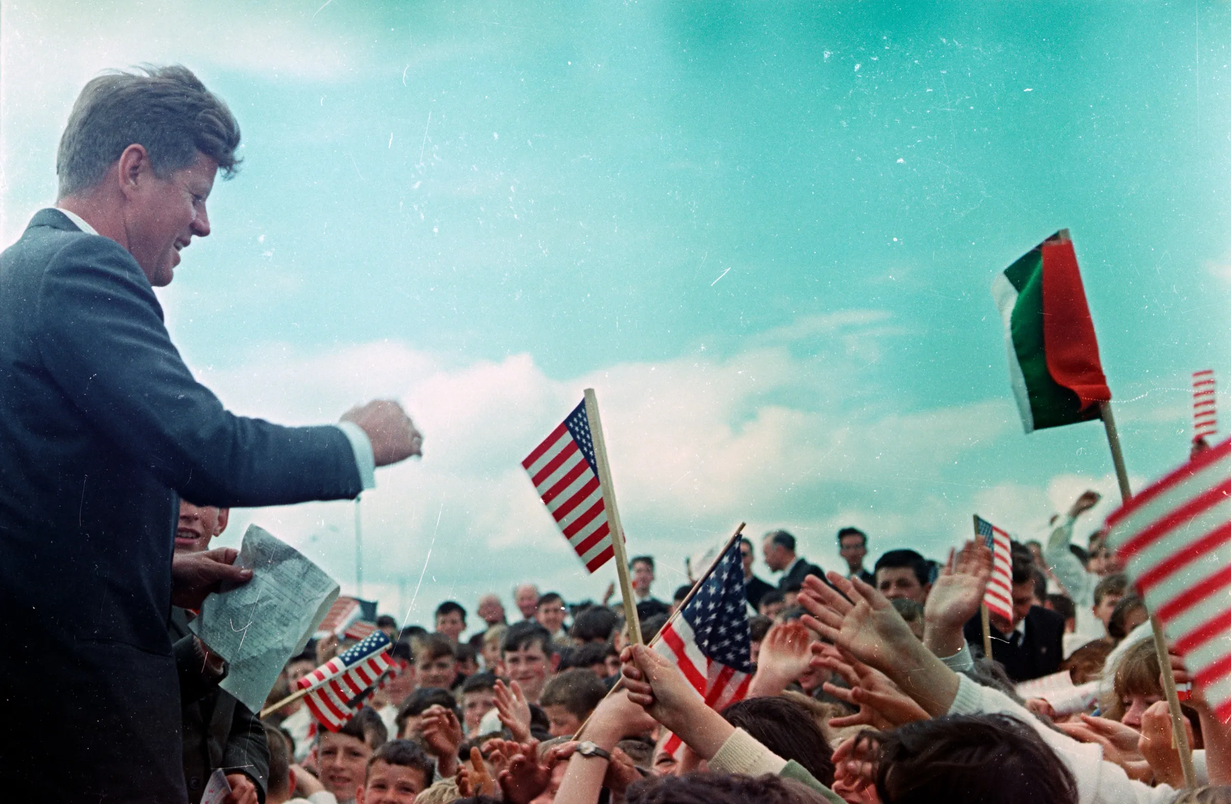 President Kennedy, smiling, reaches out to a crowd of children waving small American and Irish flags under a wide turquoise sky.