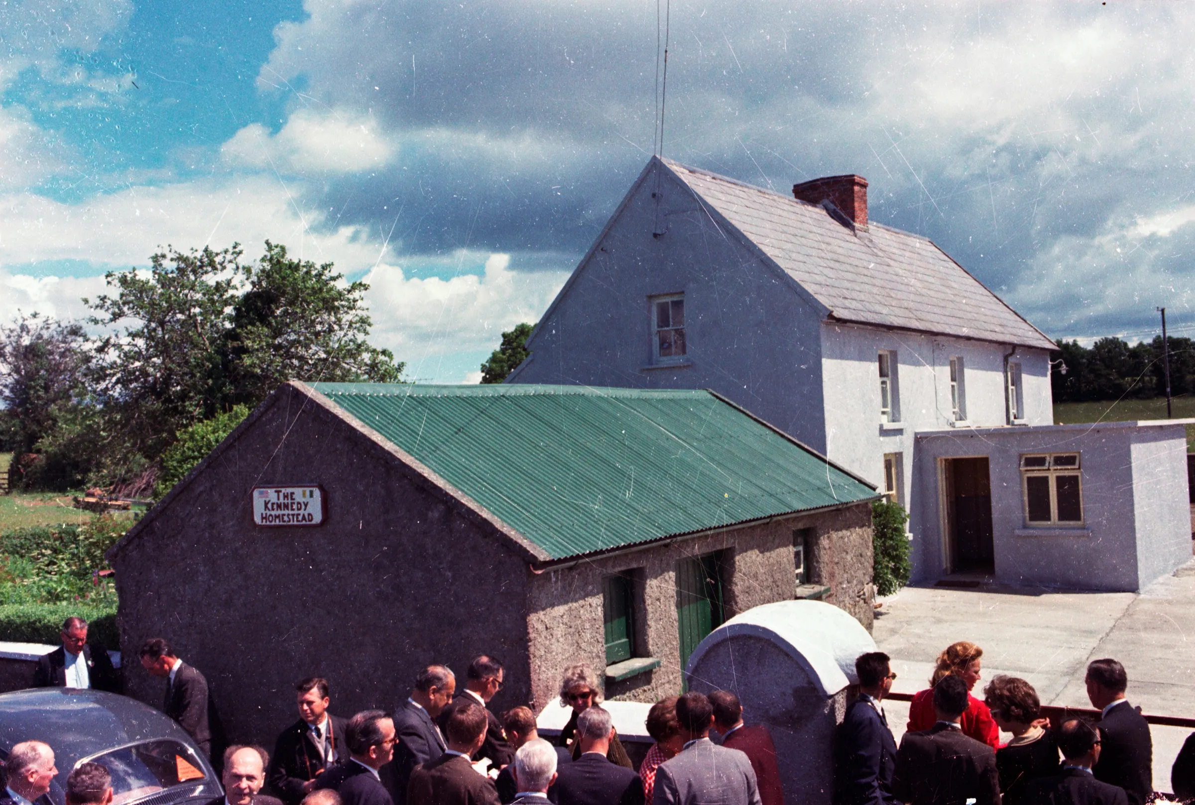 The homestead in 1963: a low stone cottage with a green corrugated roof and a hand-painted sign reading The Kennedy Homestead, a two-storey whitewashed farmhouse behind it, and men in suits gathered in the yard below a cloudy sky.