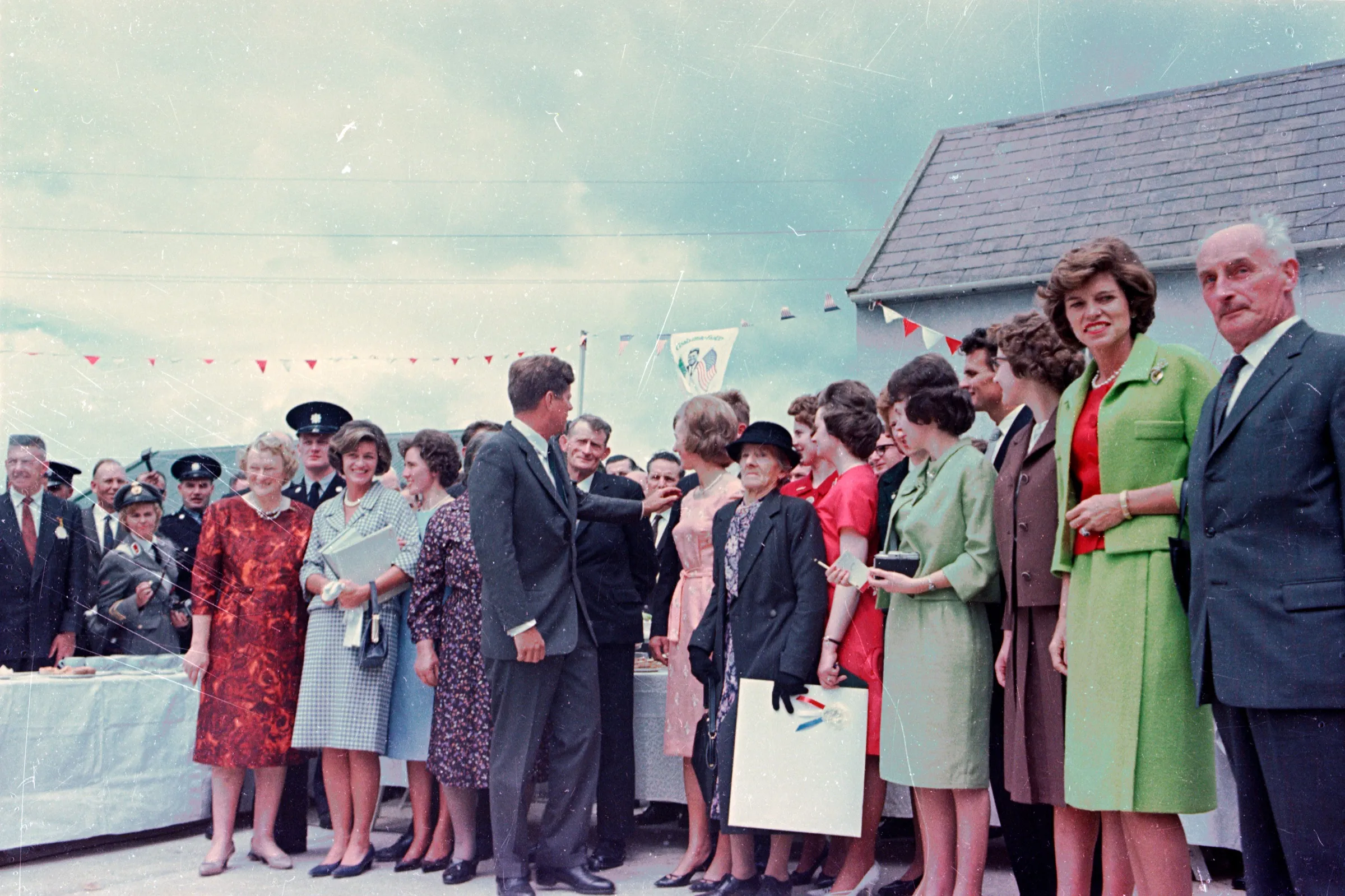 President Kennedy shaking hands in the farmyard among women in bright coats and dresses, under red and white bunting.