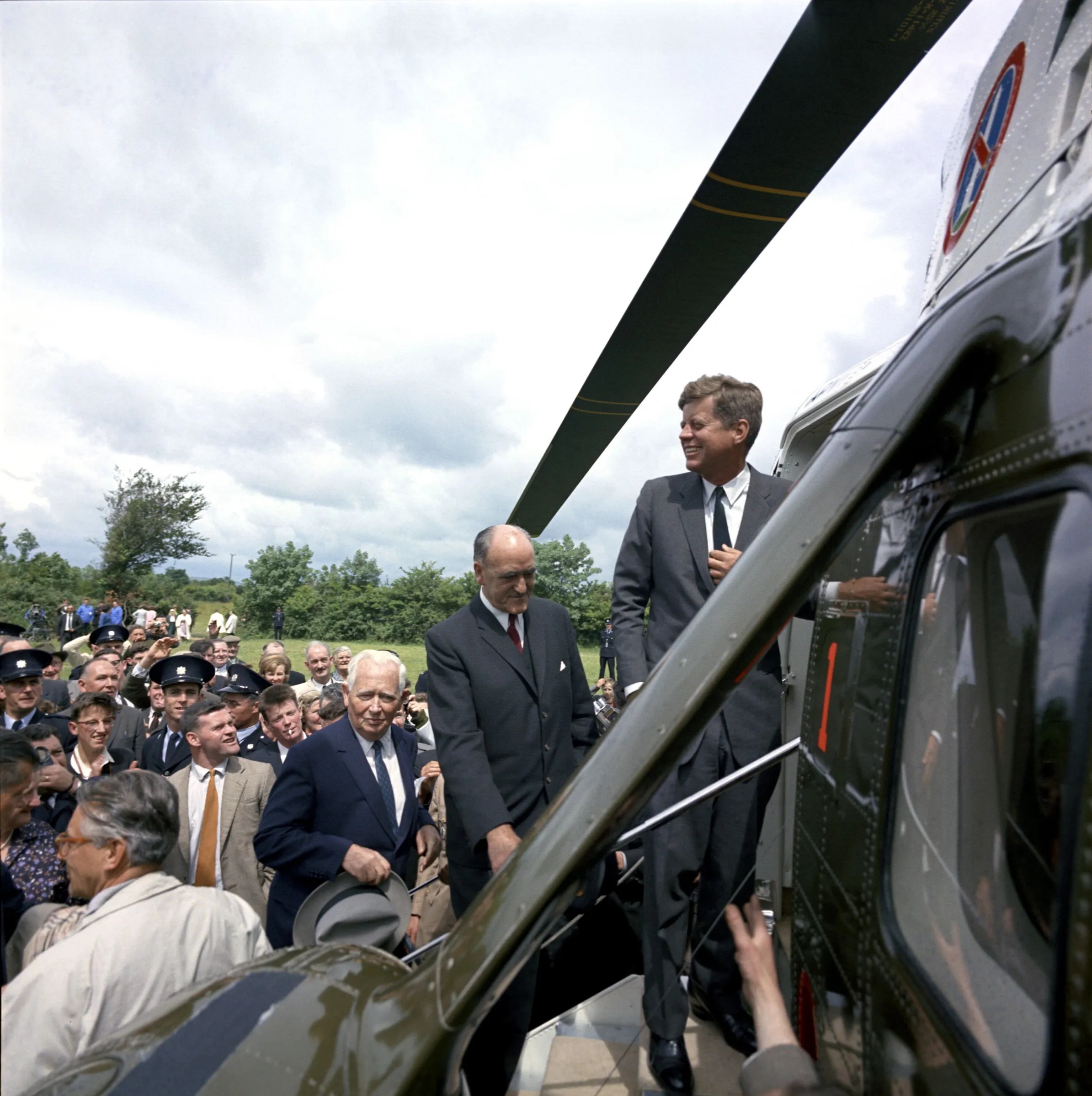 President Kennedy smiles from the steps of the Army helicopter as the crowd in the field looks on.