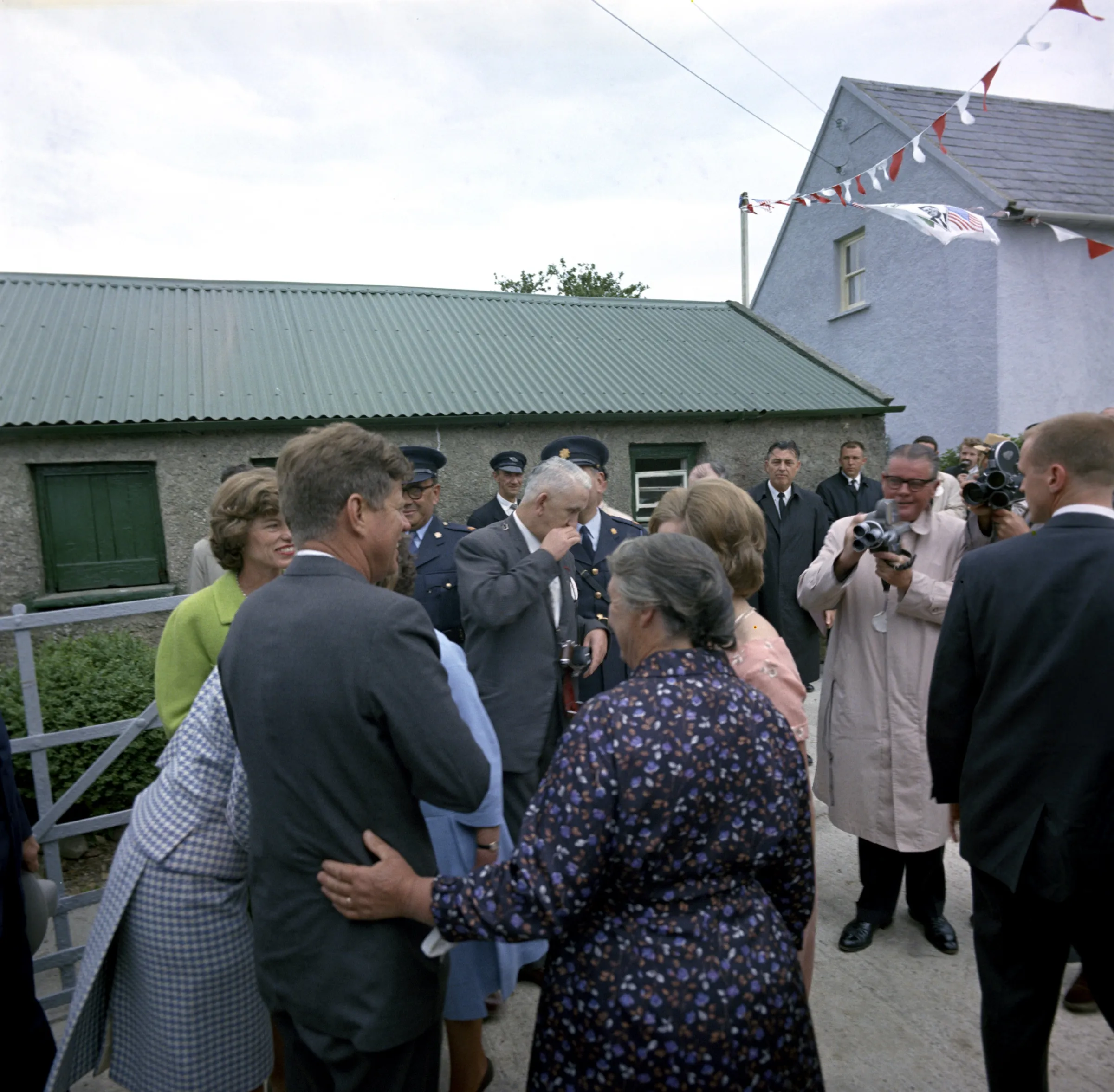 An older woman in a floral dress rests her hand on President Kennedy’s back as he greets family in the farmyard, while press photographers take pictures.