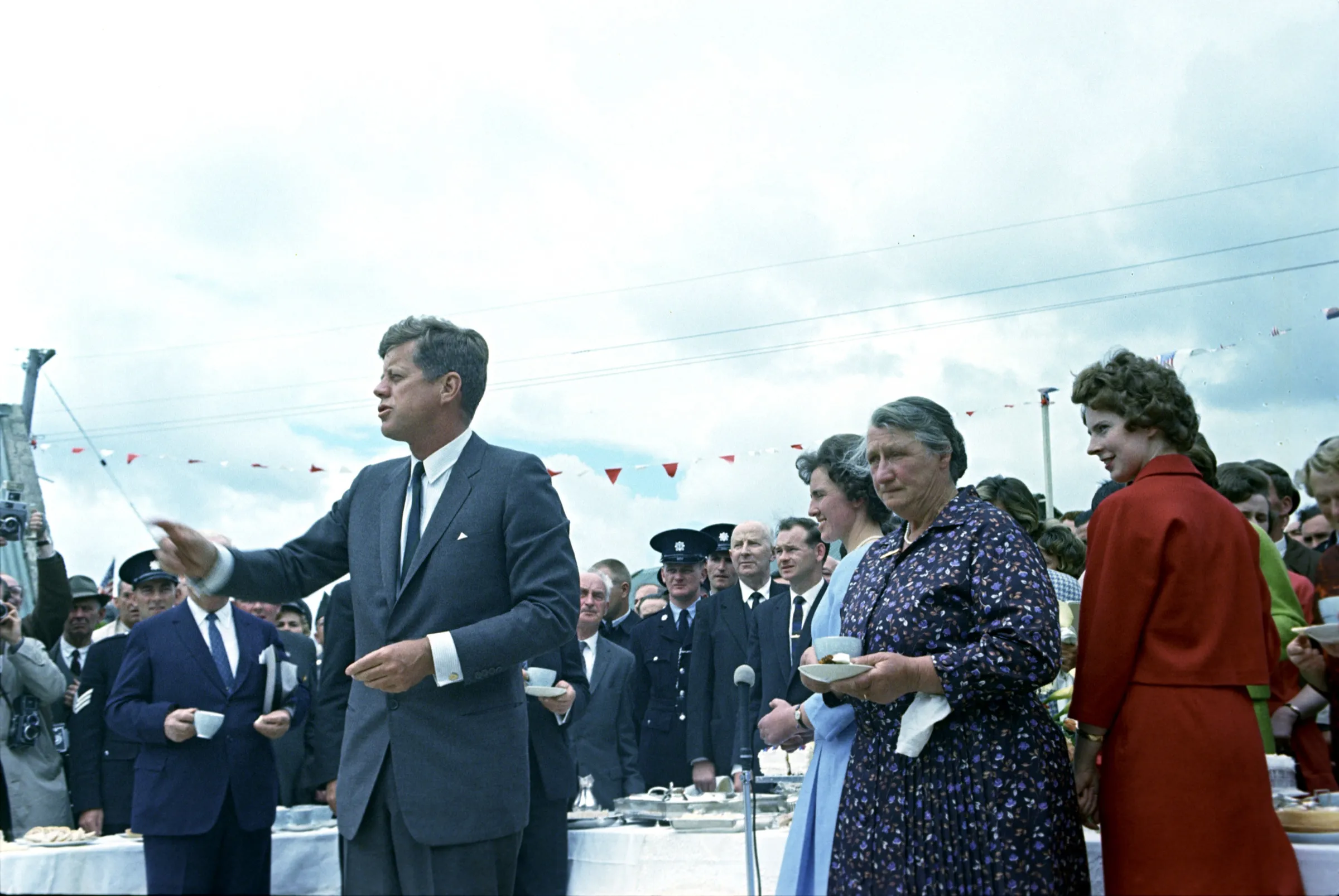 President Kennedy, in a grey suit, speaks and gestures at the reception. Beside him an older woman in a floral dress holds a cup and saucer. Tables laid for tea, bunting and uniformed officers fill the background.