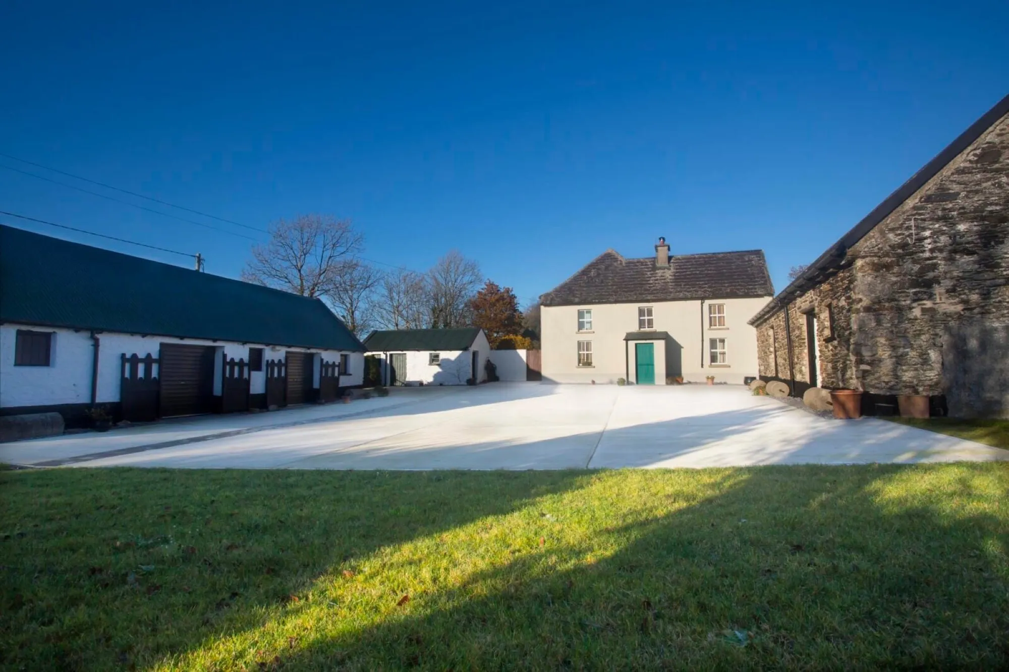 The courtyard today: a whitewashed farmhouse with a green door at the far end, a long whitewashed outbuilding on the left and a stone barn on the right, with a lawn in the foreground under a clear blue sky.
