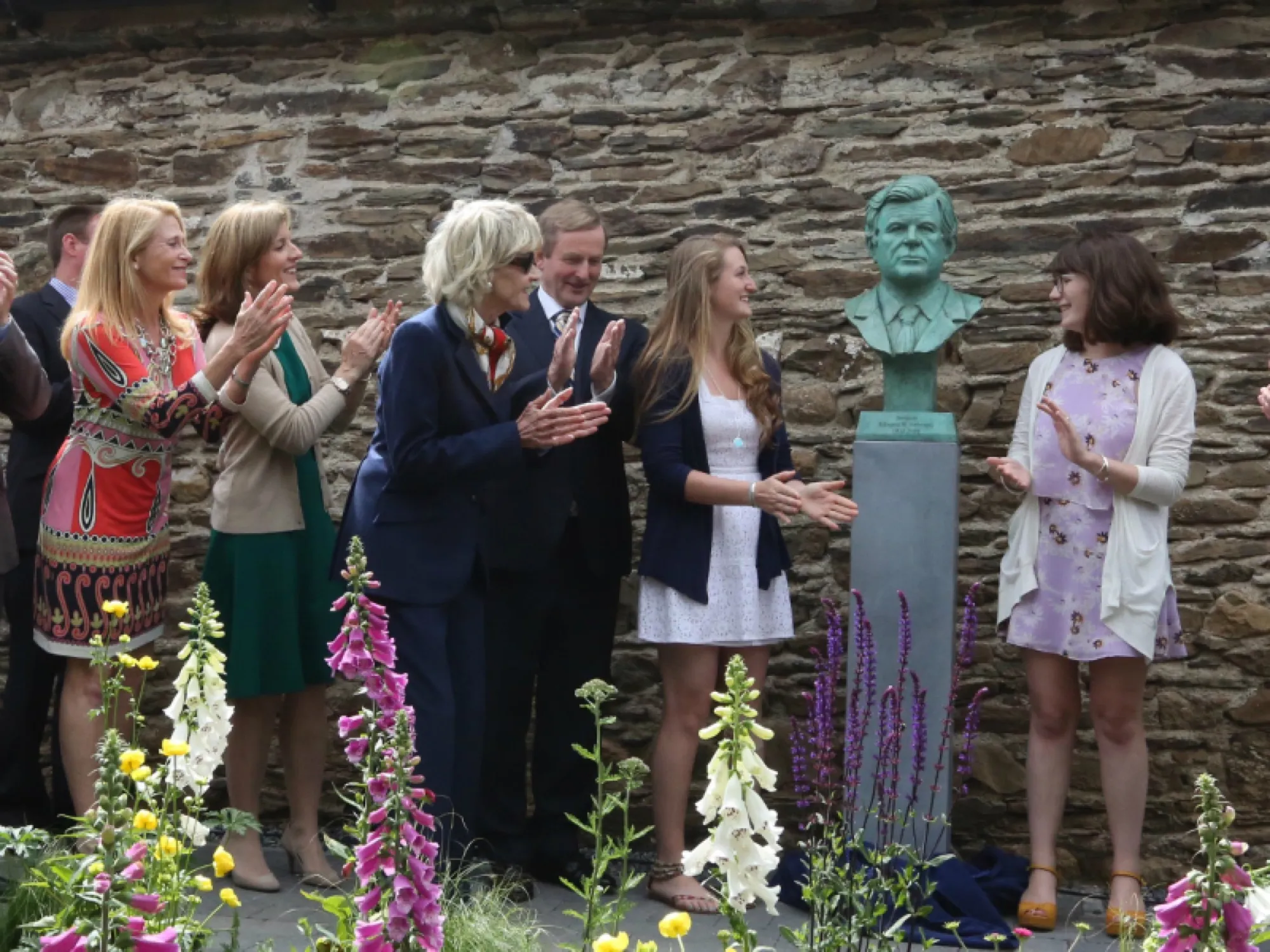 Guests applaud beside a bronze bust of Edward M. Kennedy on a stone plinth, in front of the old stone wall of the homestead, with foxgloves in the foreground.