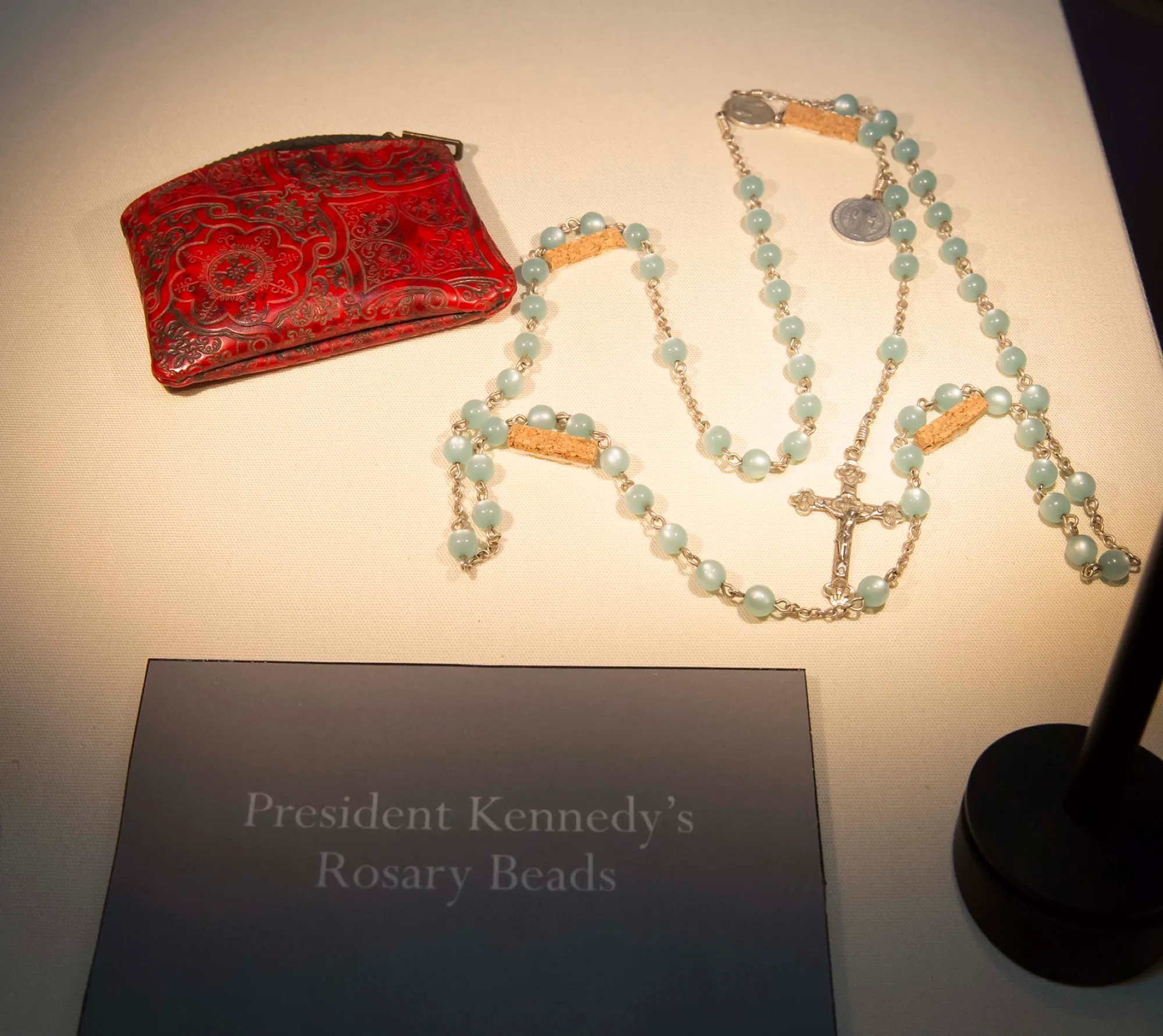A rosary of pale blue beads with a silver crucifix, beside a red brocade purse, above a dark card reading President Kennedy’s Rosary Beads.