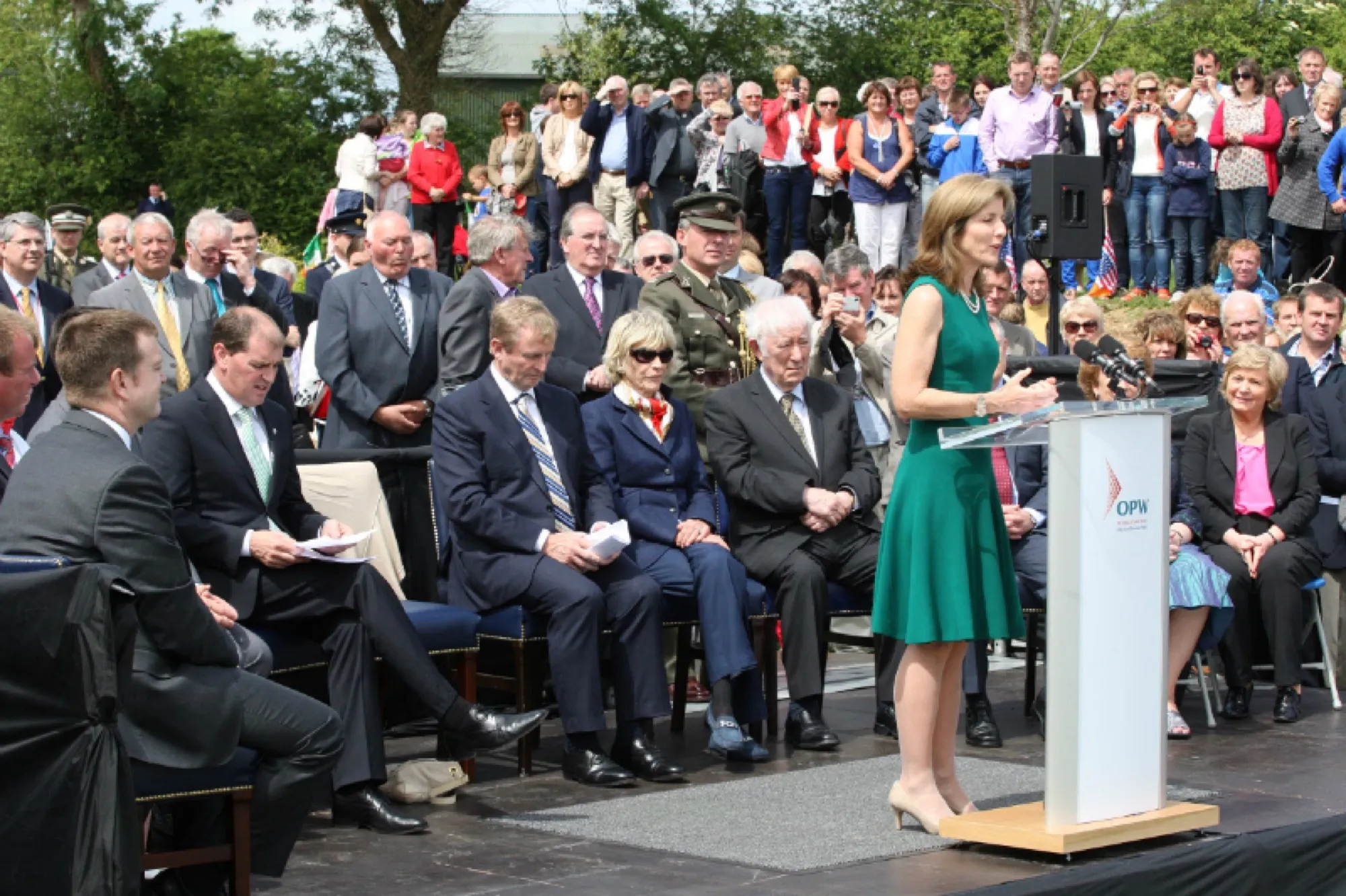 Caroline Kennedy, in a green dress, speaks from a lectern to seated guests and a large crowd at the opening of the visitor centre.