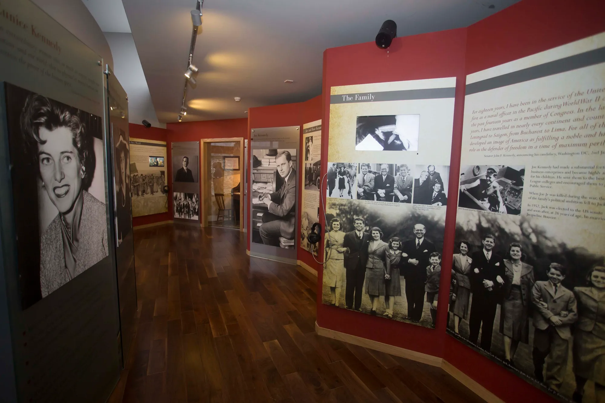 Inside the exhibition: a wooden-floored gallery lined with red and white panels of family photographs, with a portrait of a young woman in the foreground.