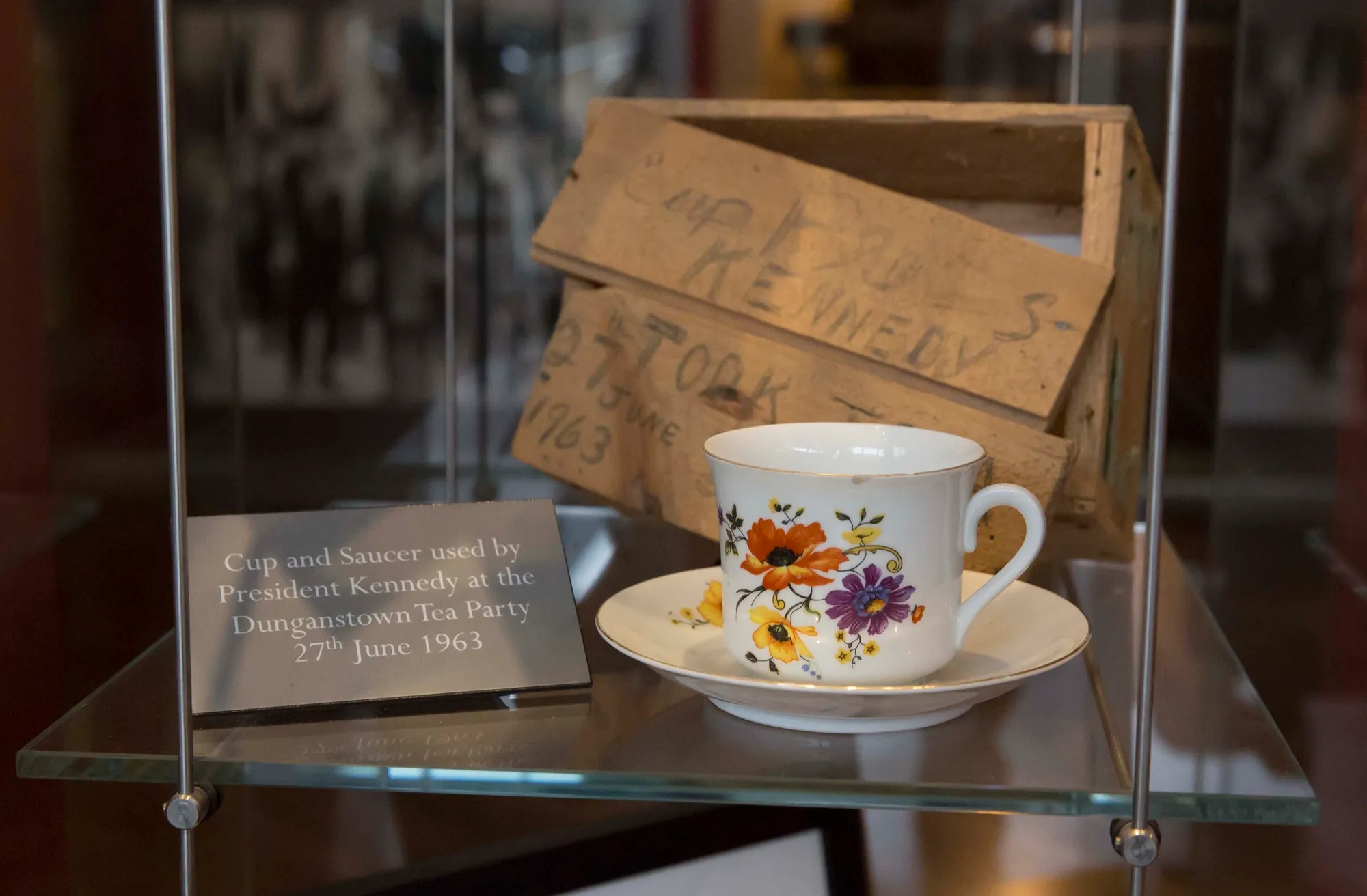 A white china cup and saucer painted with orange and purple flowers, in a glass case. A plaque beside it reads: Cup and Saucer used by President Kennedy at the Dunganstown Tea Party, 27th June 1963. Behind it stands a worn wooden box with Kennedy and 27 June 1963 written on it in pencil.